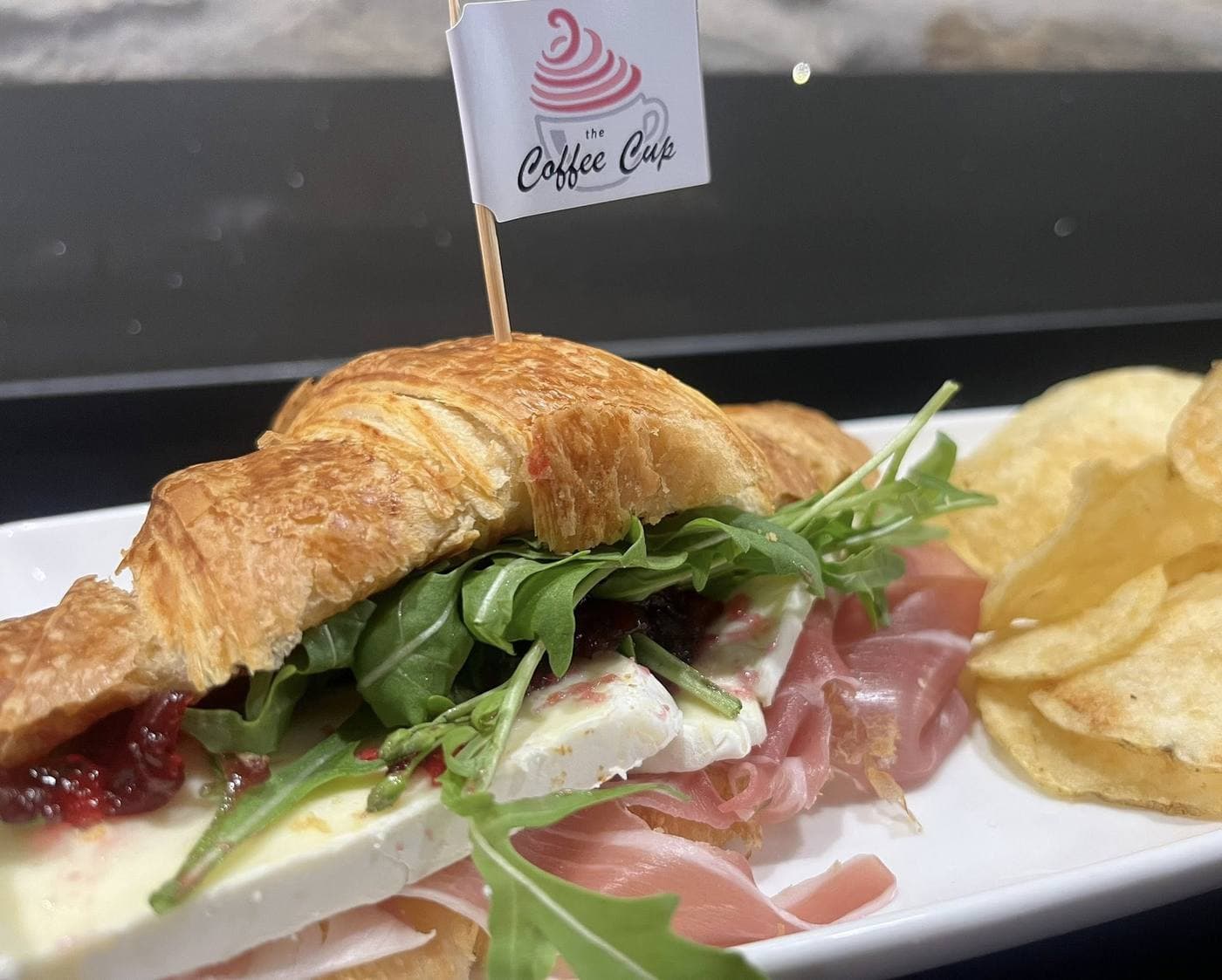 Croissant with prosciutto, brie and rocket on a white plate with kettle chips, Coffee Cup branded flag