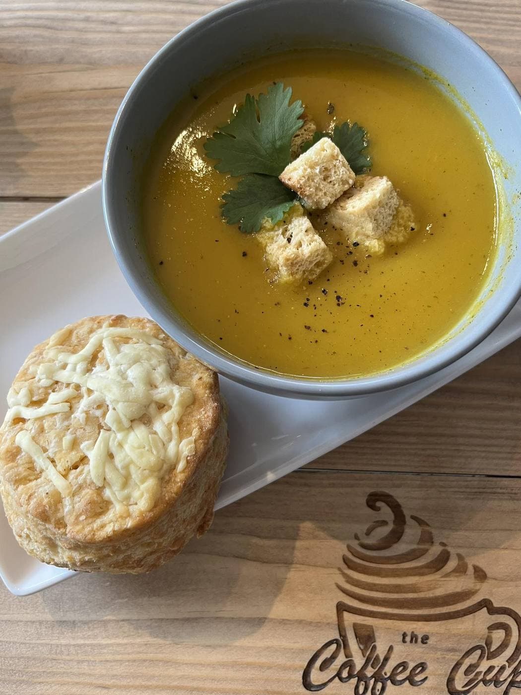 Pumpkin soup with croutons and coriander beside a cheese scone, on a board etched with the Coffee Cup logo