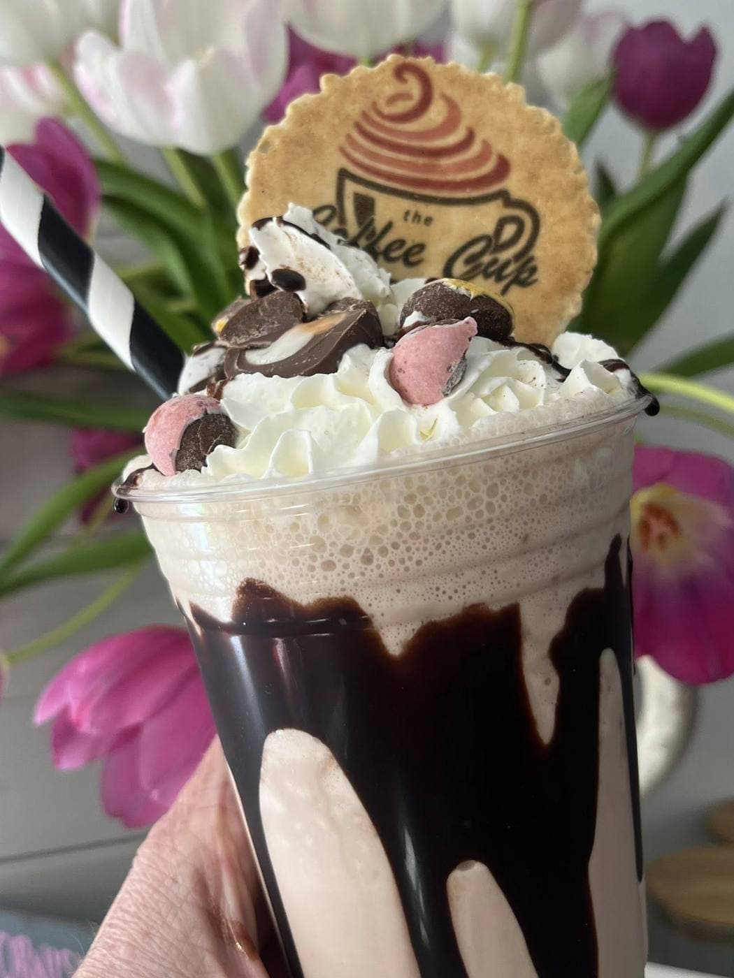 Chocolate milkshake with whipped cream, Mini Eggs and chocolate pieces, topped with a Coffee Cup branded biscuit wafer, styled against pink tulips
