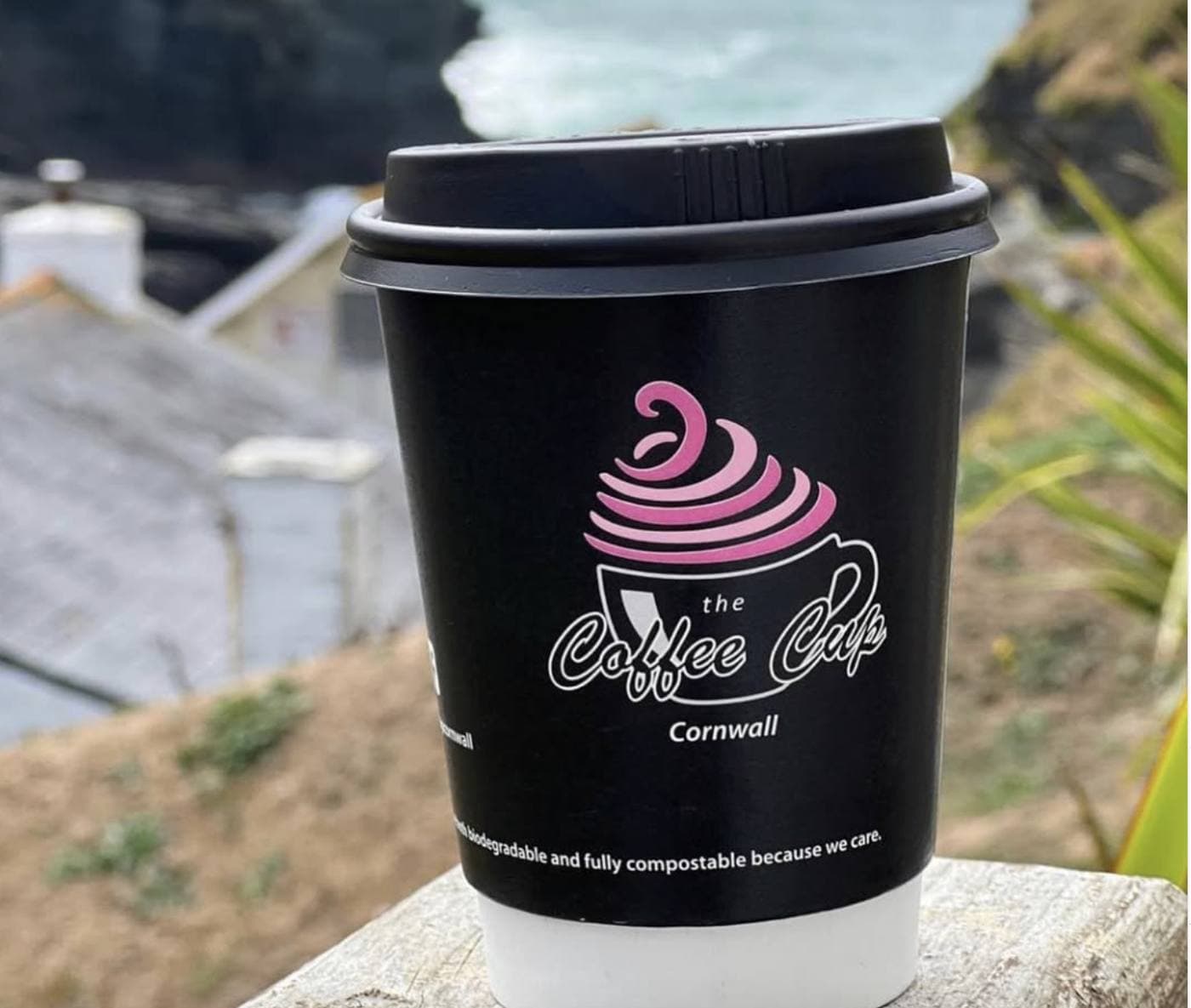 A Coffee Cup branded compostable takeaway cup on a wall overlooking the Trebarwith Strand coastline with Cornish cliffs and cottages in the background